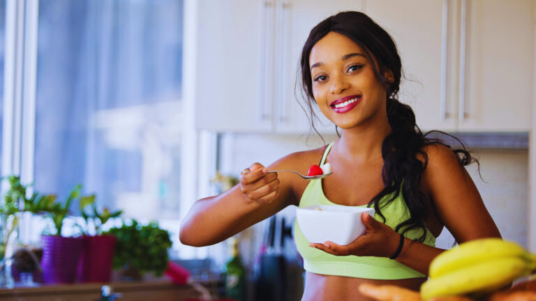 Smiling woman in sportswear holding a healthy snack in a bright kitchen, promoting smart weight loss habits.