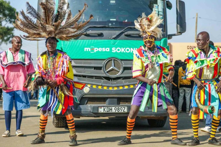 A vibrant cultural group dressed in colorful traditional attire poses in front of a green M-PESA Sokoni truck with a Mercedes-Benz logo and license plate KDH 989Z. The performers wear feathered headpieces and striped costumes, and some hold traditional musical instruments.