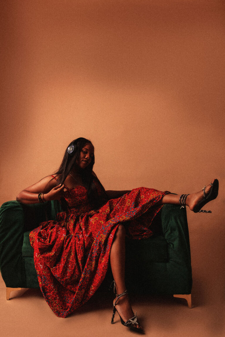 AfroReekAH seated on green couch in red patterned dress and headphones, expressing joy and introspection.