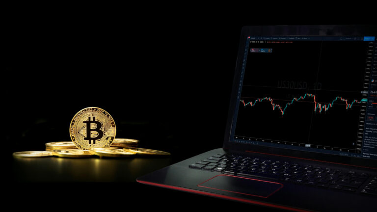 Bitcoin coins beside a laptop displaying a financial trading chart on a dark background.