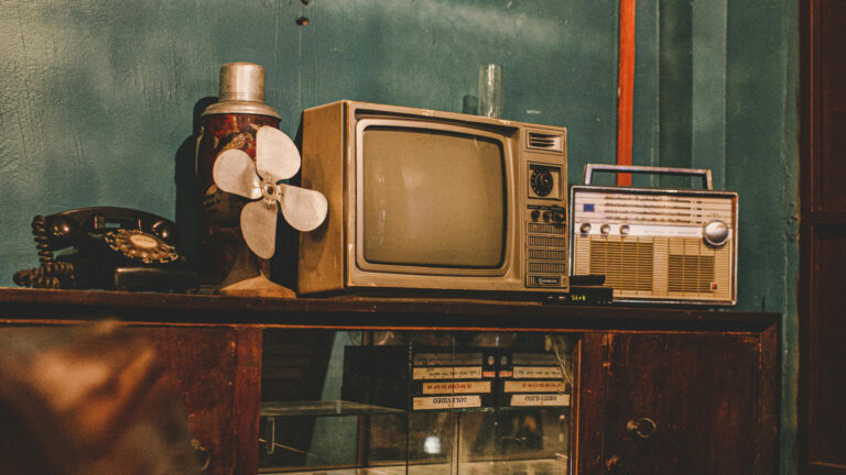 A vintage Samsung television set, a portable transistor radio, and a rotary dial telephone arranged on a wooden cabinet with glass doors, surrounded by retro household items against a teal wall.
