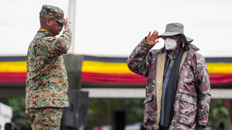 President Yoweri Museveni, wearing a face mask, scarf, and bucket hat, beside his son Gen. Muhoozi Kainerugaba, who salutes in full military uniform, with Uganda's flag colors draped behind them.