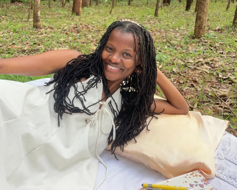 Truphine Apondi, known online as Simply Apondi, reclining on a blanket in a wooded grove, smiling at the camera with long braided locs and a beaded headband, wearing a white sleeveless dress.