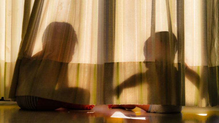 Two children silhouette playfully sitting behind sheer curtains in sunlight.