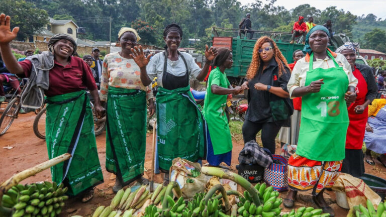 A vibrant group of Kenyan women market traders smiling and celebrating alongside Safaricom representatives during an M-PESA Sokoni activation in Webuye, featuring fresh green bananas in the foreground.
