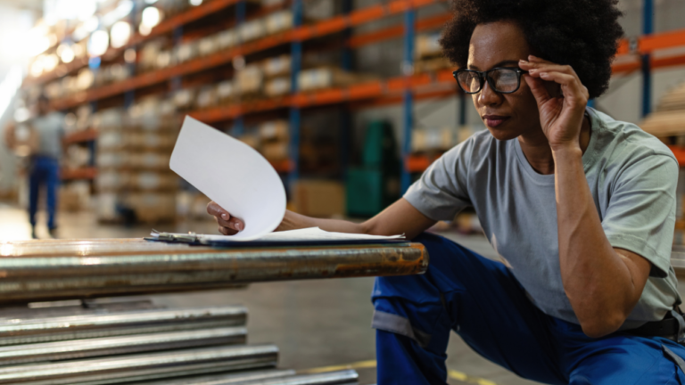 Woman in a warehouse checking metal equipment stock against paperwork on a clipboard