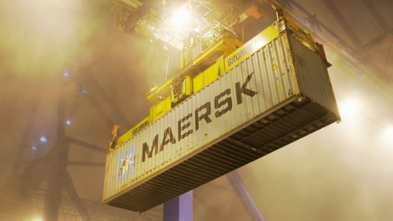 A Maersk shipping container lifted by a crane at a port terminal at night, surrounded by fog and floodlights.