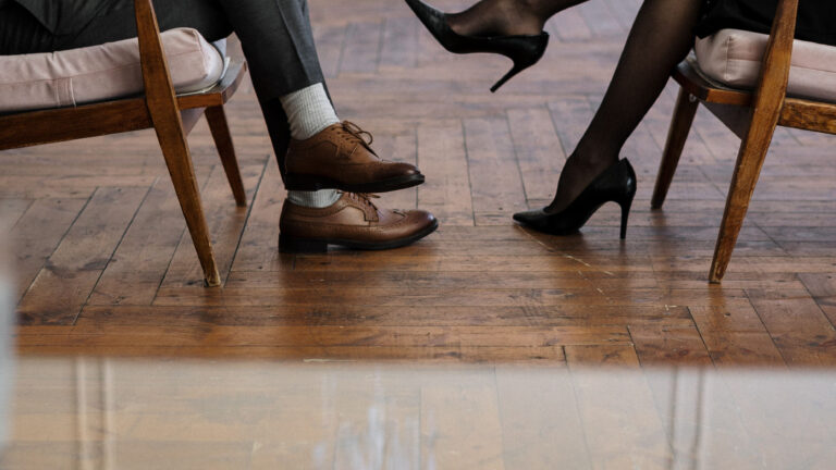 Two people seated across from each other in armchair chairs, showing only their legs and formal footwear during a serious conversation or therapy session.