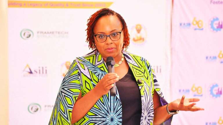Ms. Winnie Ngumi speaking with a microphone in front of branded event backdrops featuring KAM (Kenya Association of Manufacturers) logos, Frametech Structures, and Asili Roofing Tiles.
