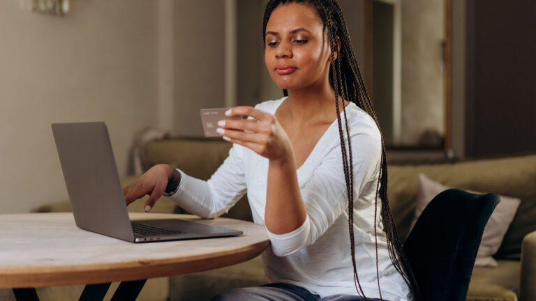 A woman with braided hair sits at a table, holding a card up to read while her other hand rests on a laptop keyboard.