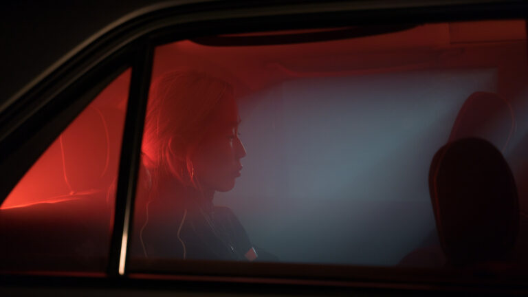 Woman with hoop earrings sitting quietly in the back seat of a car at night, lit by red light through the window, gazing away from the camera