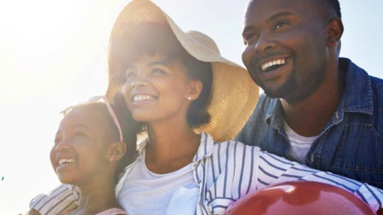 A father, mother and daughter smile at the beach, holding a beach ball, backlit by sunlight