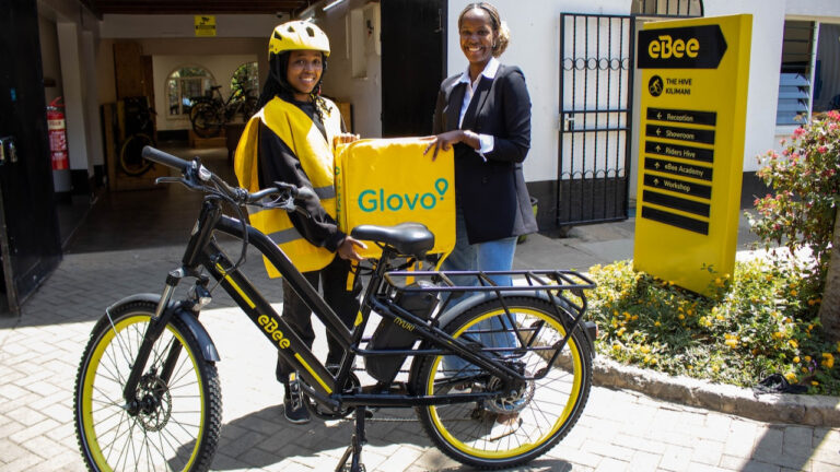 Liz Wambua of Glovo Kenya hands a yellow Glovo delivery bag to rider Yvonne Muribi beside an electric bicycle at the eBee Hive in Kilimani.