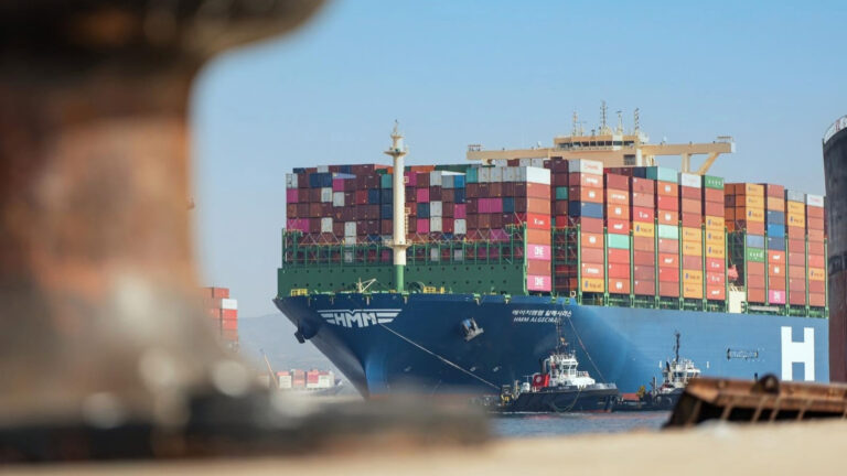 HMM container ship Algeciras stacked with containers, guided by two tugboats, seen past a blurred mooring bollard.