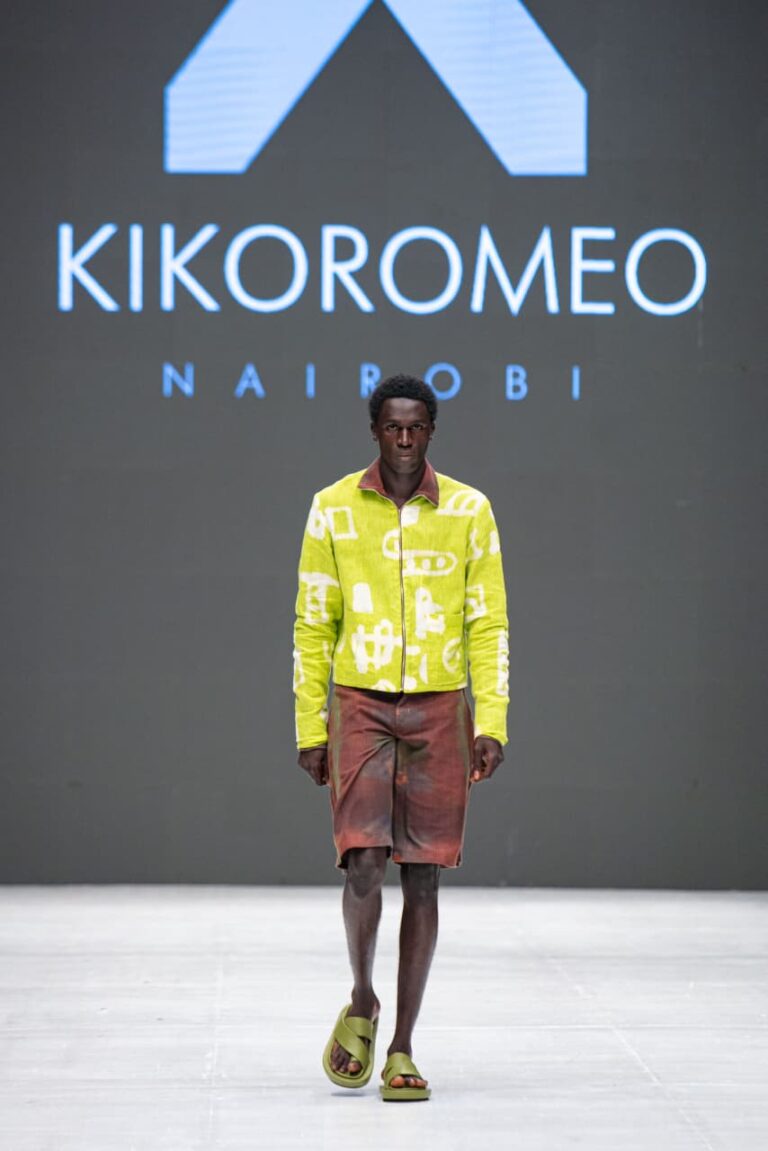 Model walks a runway in a lilac patterned long coat and matching trousers in front of a KikoRomeo Nairobi sign