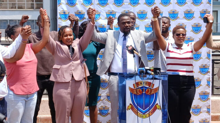 KMPDU Secretary General Davji Atellah speaks at a podium during a press briefing, surrounded by union members raising joined hands in a show of solidarity.