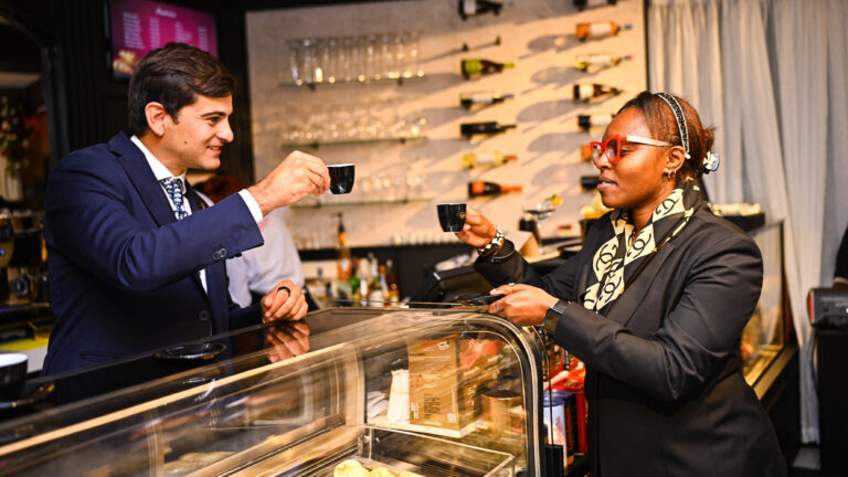 Two people raise small espresso cups across a glass pastry counter inside Café Barbera, Nairobi.