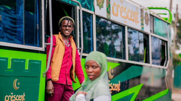 A green matatu with gold “Crescent” lettering. A man in a beanie, red shirt and orange vest stands in the open doorway, and a woman in a pale green hijab is in the foreground.