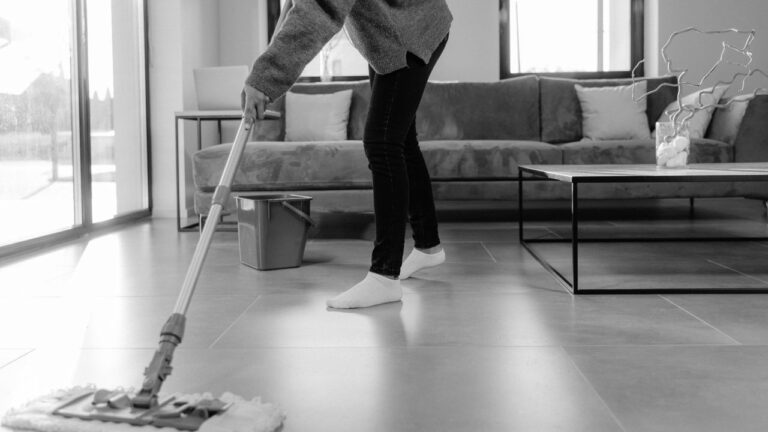 Person mopping a tiled living room floor beside a sofa, coffee table and mop bucket, in black and white