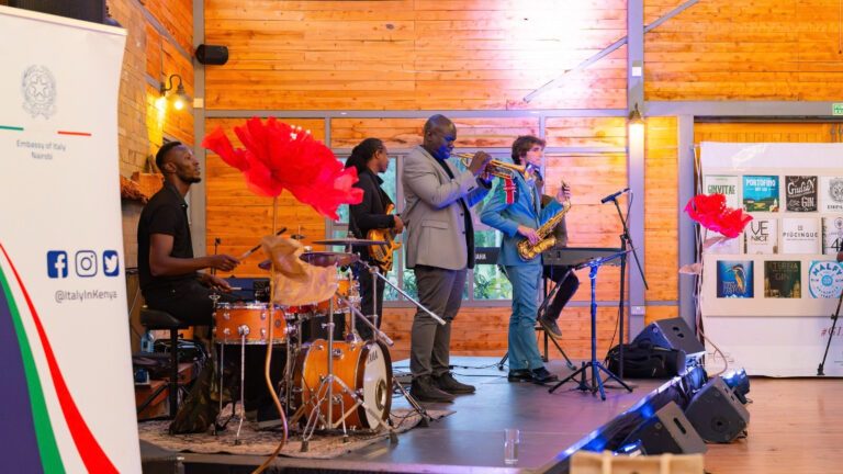 Four musicians on a stage at Shamba Events: a drummer, a bass guitarist, a trumpeter and a saxophonist. An Embassy of Italy Nairobi banner stands at left and a display of Italian gin labels hangs at right.