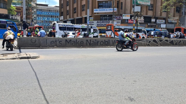Buses and motorcycles on a Nairobi street beside a median painted with a city slogan