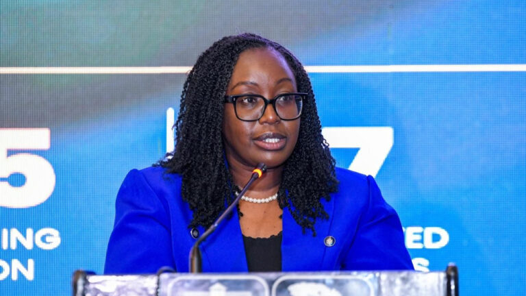 Risper Ohaga, Group Chief Executive of APA Apollo Group, speaks at a podium wearing a blue blazer, glasses and a pearl necklace
